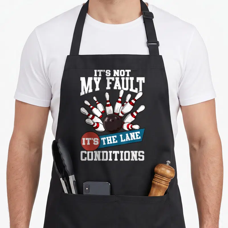 Fault bowling life apron - Cartoon bowling ball hitting pins with It's Not My Fault text — shown on a black cotton canvas apron against a white background