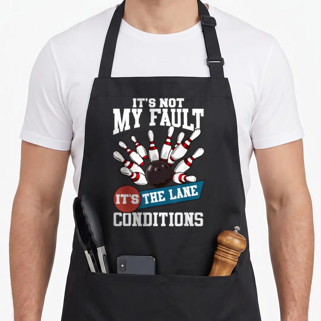 Fault bowling life apron - Cartoon bowling ball hitting pins with It's Not My Fault text — shown on a black cotton canvas apron against a white background
