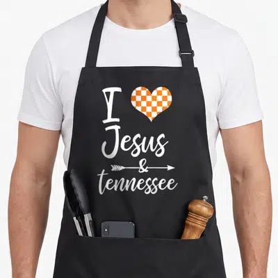 Jesus tennessee apron - Stacked white text I Jesus & tennessee with orange checkerboard heart and arrow through — shown on a black cotton canvas apron against a white background