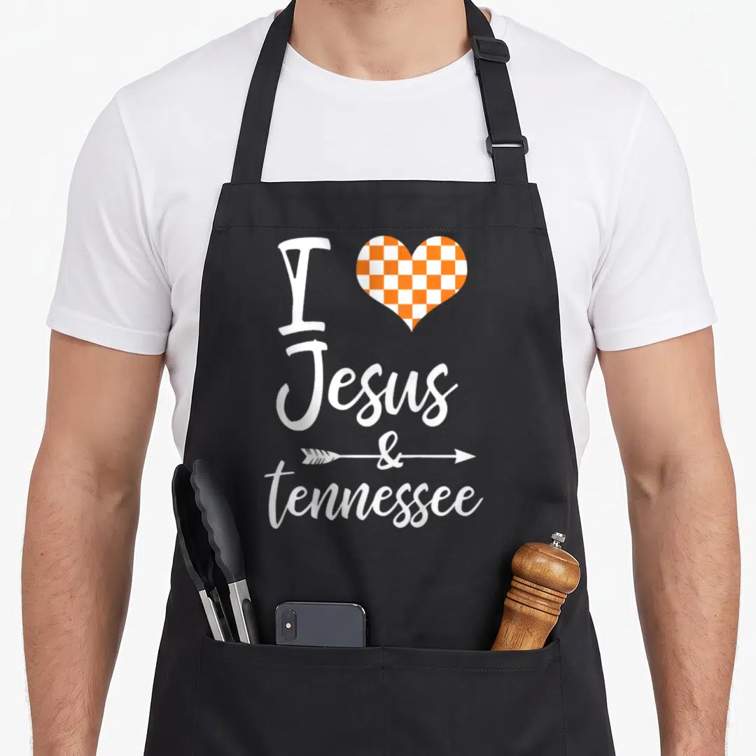 Jesus tennessee apron - Stacked white text I Jesus & tennessee with orange checkerboard heart and arrow through — shown on a black cotton canvas apron against a white background