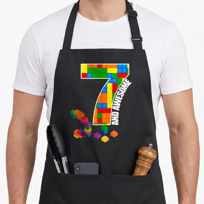 Block brick building aprons - Number 7 filled with multicolored toy bricks and AND AWESOME banner text — shown on a black cotton canvas apron against a white background