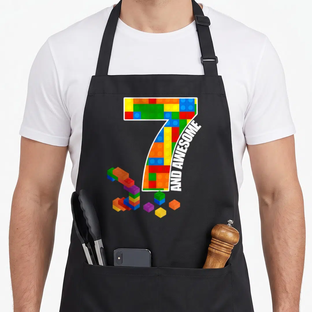 Block brick building aprons - Number 7 filled with multicolored toy bricks and AND AWESOME banner text — shown on a black cotton canvas apron against a white background