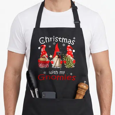 Funny family christmas gnome apron - Three cartoon gnomes with red hats holding gifts, camera, and tree under arched — shown on a black cotton canvas apron against a white background