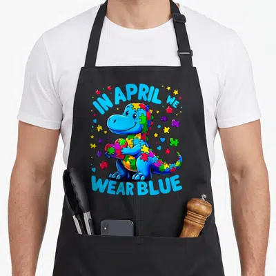 Blue april apron - Cartoon dinosaur holding puzzle heart with IN APRIL WE WEAR BLUE text — shown on a black cotton canvas apron against a white background