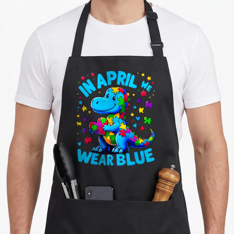 Blue april apron - Cartoon dinosaur holding puzzle heart with IN APRIL WE WEAR BLUE text — shown on a black cotton canvas apron against a white background