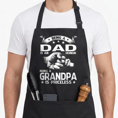 Honor grandpa apron - White distressed text Being A Dad Is An Honor Being A Grandpa Is Priceless with hands clasping — shown on a black cotton canvas apron against a white background