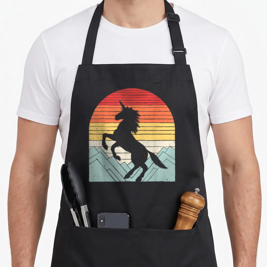 Catch unicorn apron - Grey unicorn silhouette on striped red orange yellow sun with light blue mountains — shown on a black cotton canvas apron against a white background