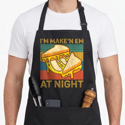 Makin grilled apron - Cartoon grilled cheese over yellow red blue and green stripes with text I'M MAKE'N EM AT NIGHT — shown on a black cotton canvas apron against a white background