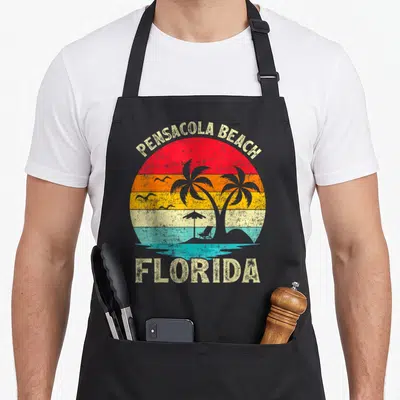 Florida family aprons - Retro striped sun with PENSACOLA BEACH FLORIDA text, palm trees, birds, and beach chair — shown on a black cotton canvas apron against a white background