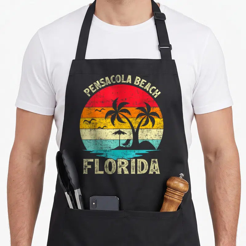 Florida family aprons - Retro striped sun with PENSACOLA BEACH FLORIDA text, palm trees, birds, and beach chair — shown on a black cotton canvas apron against a white background