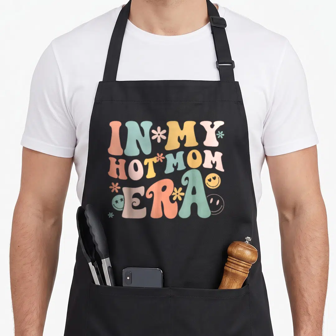 Hot mother's day aprons - Stacked retro text In My Hot Mom Era with peach blue yellow pink green flowers and smiley — shown on a black cotton canvas apron against a white background