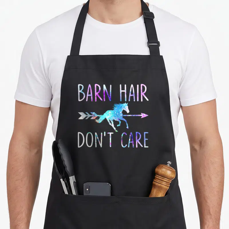 Barn hair apron - Unicorn overlapping arrow with BARN HAIR DON'T CARE text in purple, blue, pink, and white — shown on a black cotton canvas apron against a white background