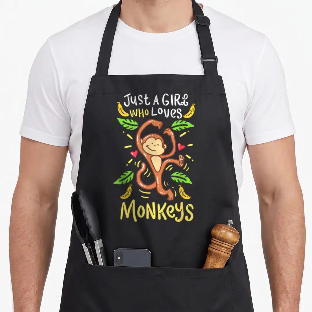 Monkey zookeeper apron - Cartoon brown monkey with bananas, green leaves, and Just A Girl Who Loves Monkeys text — shown on a black cotton canvas apron against a white background