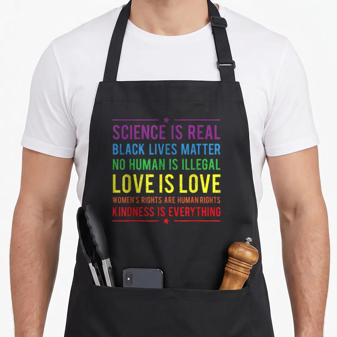 Kindness science apron - Rainbow stacked text reading Science is Real Black Lives Matter No Human is Illegal Love is — shown on a black cotton canvas apron against a white background