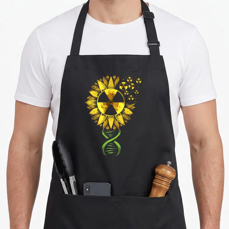 Nuclear x ray apron - Yellow sunflower with radiation center and green DNA helix stem on black background — shown on a black cotton canvas apron against a white background