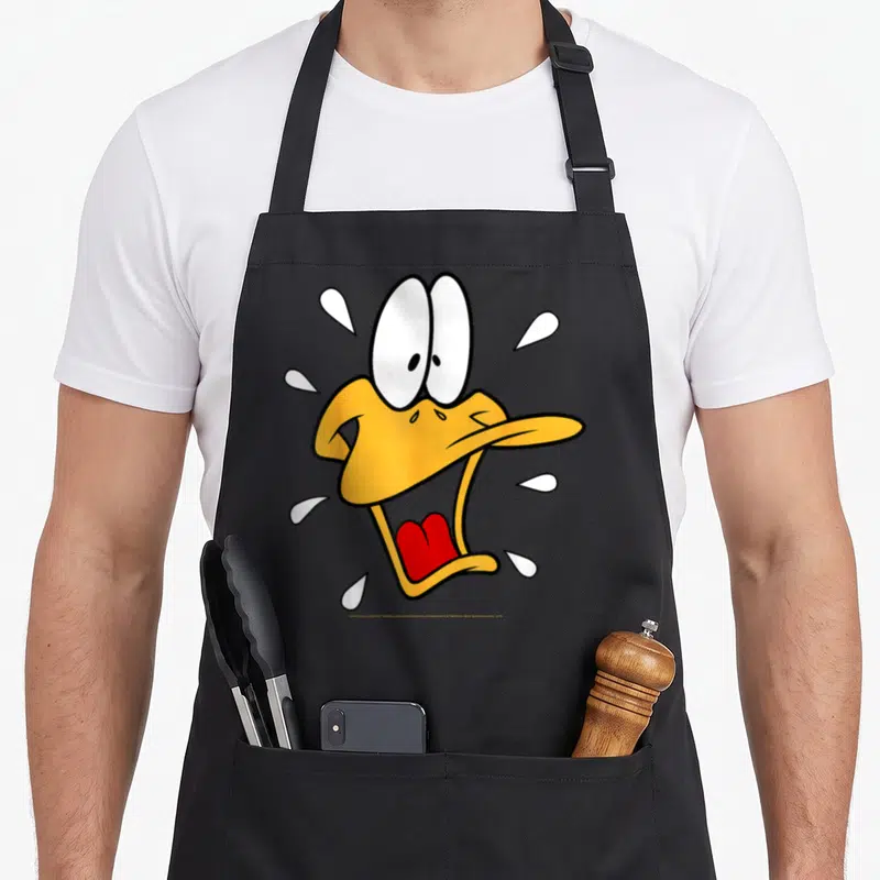 Surprised duck apron - Cartoon Daffy Duck head with white sweat drops and red tongue — shown on a black cotton canvas apron against a white background