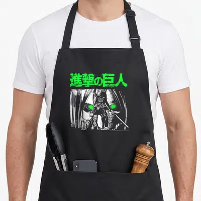 Eye anime apron - Green Japanese text above small character standing before large face with glowing green eyes — shown on a black cotton canvas apron against a white background