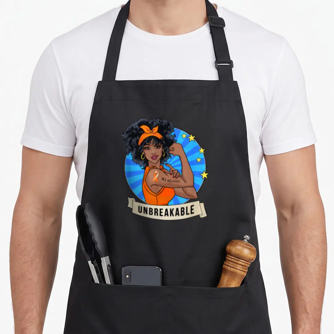 Unbreakable manuscript apron - Cartoon woman flexing arm with orange ribbon and MS Warrior text on blue sunburst — shown on a black cotton canvas apron against a white background