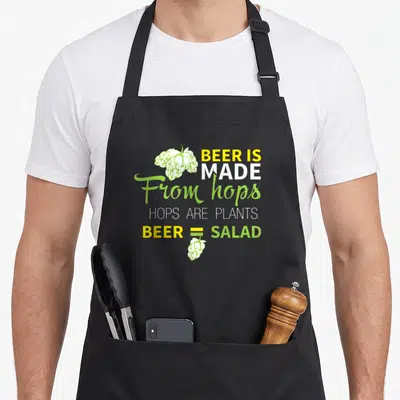 Hop beer apron - Stacked text reading beer equals salad with green and white hop clusters — shown on a black cotton canvas apron against a white background