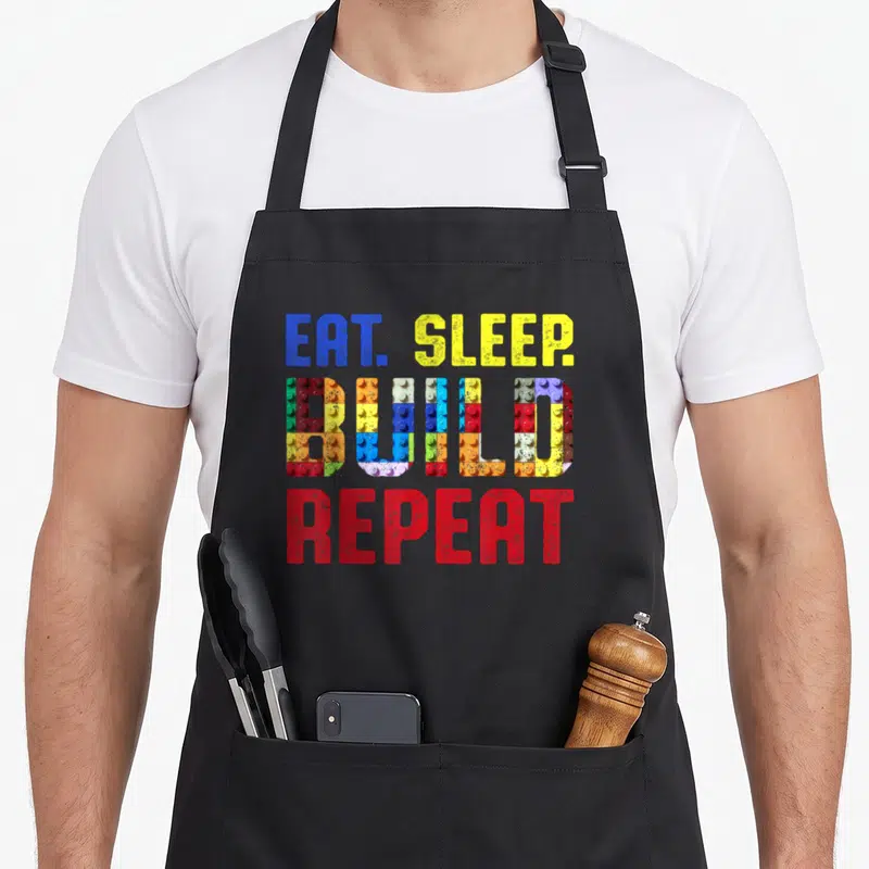Sleep building apron - EAT SLEEP BUILD REPEAT text with colorful brick pattern fill in blue yellow red green — shown on a black cotton canvas apron against a white background