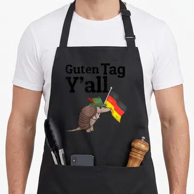 Guten texas flag apron - Cartoon armadillo in green hat holding black red yellow flag under Guten Tag Y'all text — shown on a black cotton canvas apron against a white background