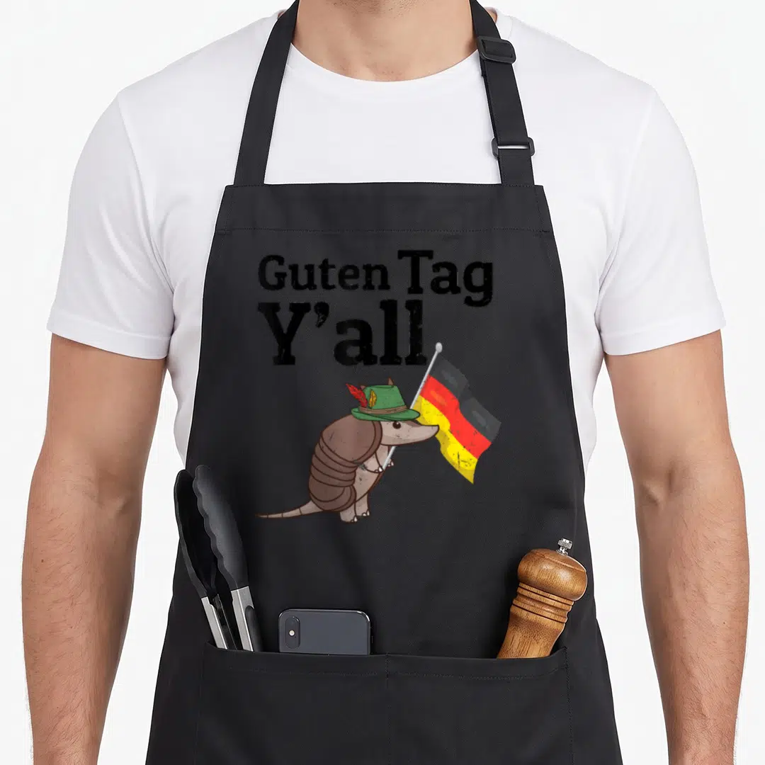 Guten texas flag apron - Cartoon armadillo in green hat holding black red yellow flag under Guten Tag Y'all text — shown on a black cotton canvas apron against a white background