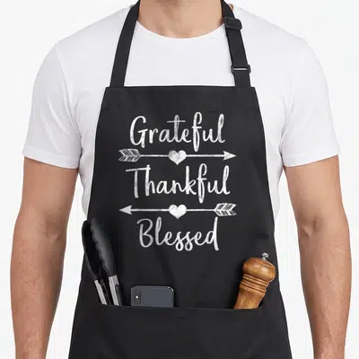 Arrow grateful thanksgiving apron - White script text Grateful Thankful Blessed separated by arrows with hearts — shown on a black cotton canvas apron against a white background
