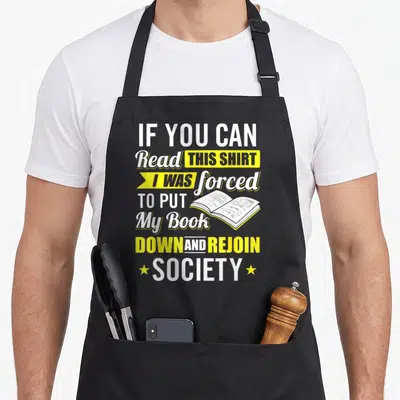 Forced funny book apron - Stacked white and yellow text with open book illustration and two stars — shown on a black cotton canvas apron against a white background