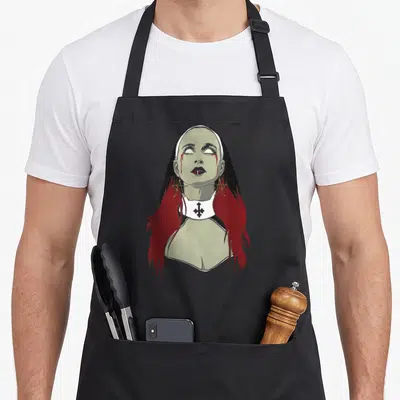 Unholy goth apron - Cartoon nun with red hair, blood tears, inverted cross collar, and pentagram earrings — shown on a black cotton canvas apron against a white background