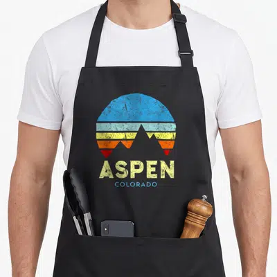 Funny aspen colorado apron - Retro sunburst with blue yellow orange red grey stripes and grey mountain silhouettes — shown on a black cotton canvas apron against a white background