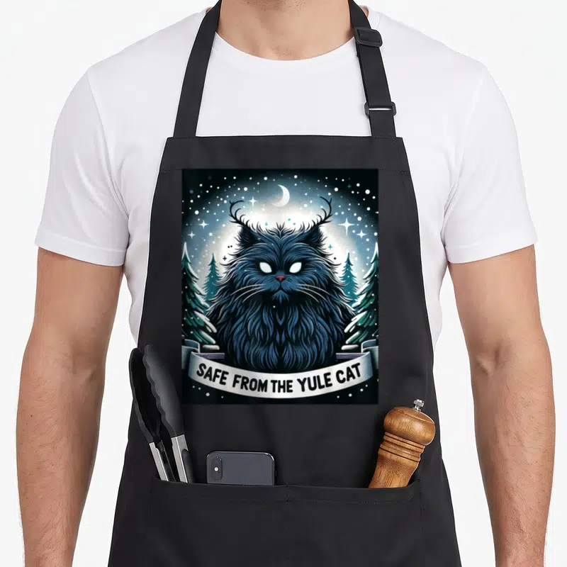 Safe christmas cat apron - Fluffy dark blue horned cat under moon and stars with banner reading SAFE FROM THE YULE CAT — shown on a black cotton canvas apron against a white background