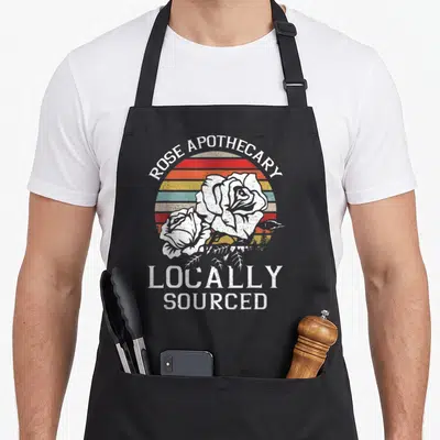 Rose pharmacist apron - Retro graphic of two white roses over a striped sun with Rose Apothecary Locally Sourced text — shown on a black cotton canvas apron against a white background