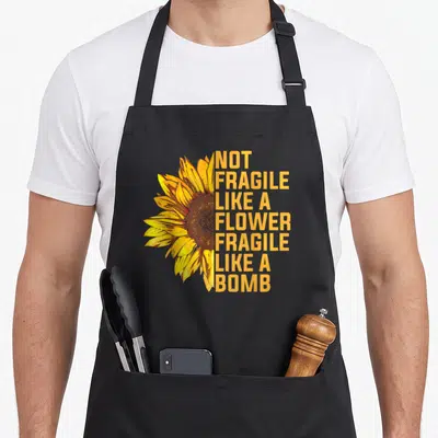 Fragile sunflower apron - Realistic sunflower with stacked orange text reading NOT FRAGILE LIKE A FLOWER FRAGILE LIKE — shown on a black cotton canvas apron against a white background