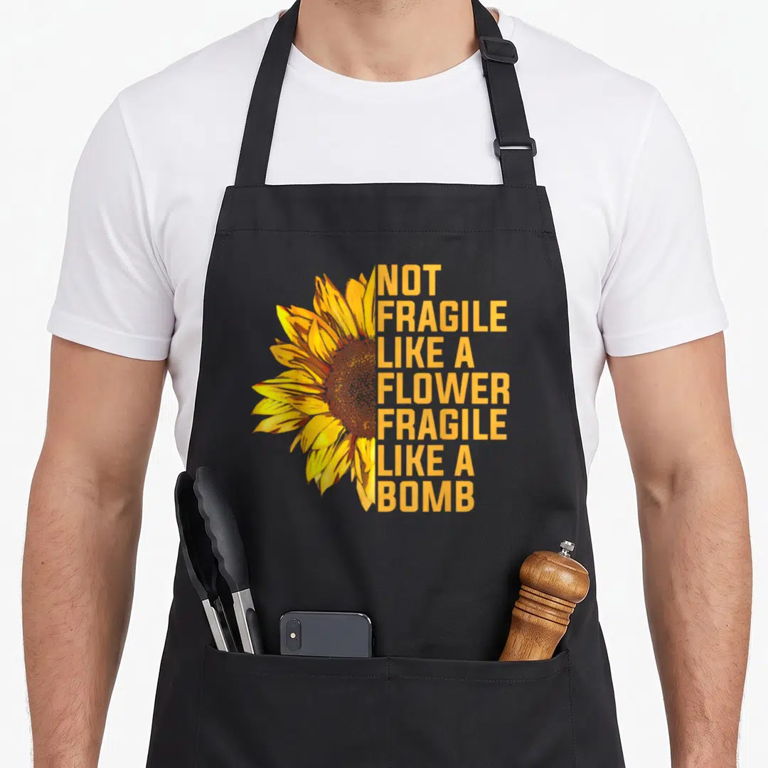 Fragile sunflower apron - Realistic sunflower with stacked orange text reading NOT FRAGILE LIKE A FLOWER FRAGILE LIKE — shown on a black cotton canvas apron against a white background