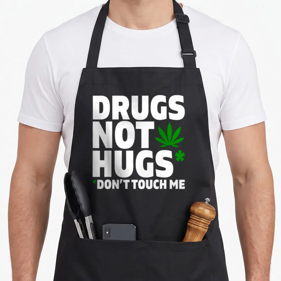 Drug smoker apron - White stacked text DRUGS NOT HUGS with green cannabis leaf and asterisks — shown on a black cotton canvas apron against a white background