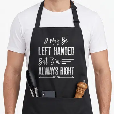 May left apron - White stacked text I May Be Left Handed But Always Right with double-headed chevron arrow below — shown on a black cotton canvas apron against a white background
