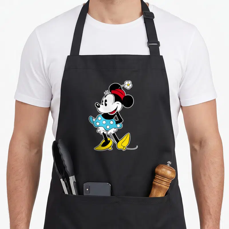 Flower mouse mom apron - Minnie Mouse standing in blue dress with white flower on red headband — shown on a black cotton canvas apron against a white background