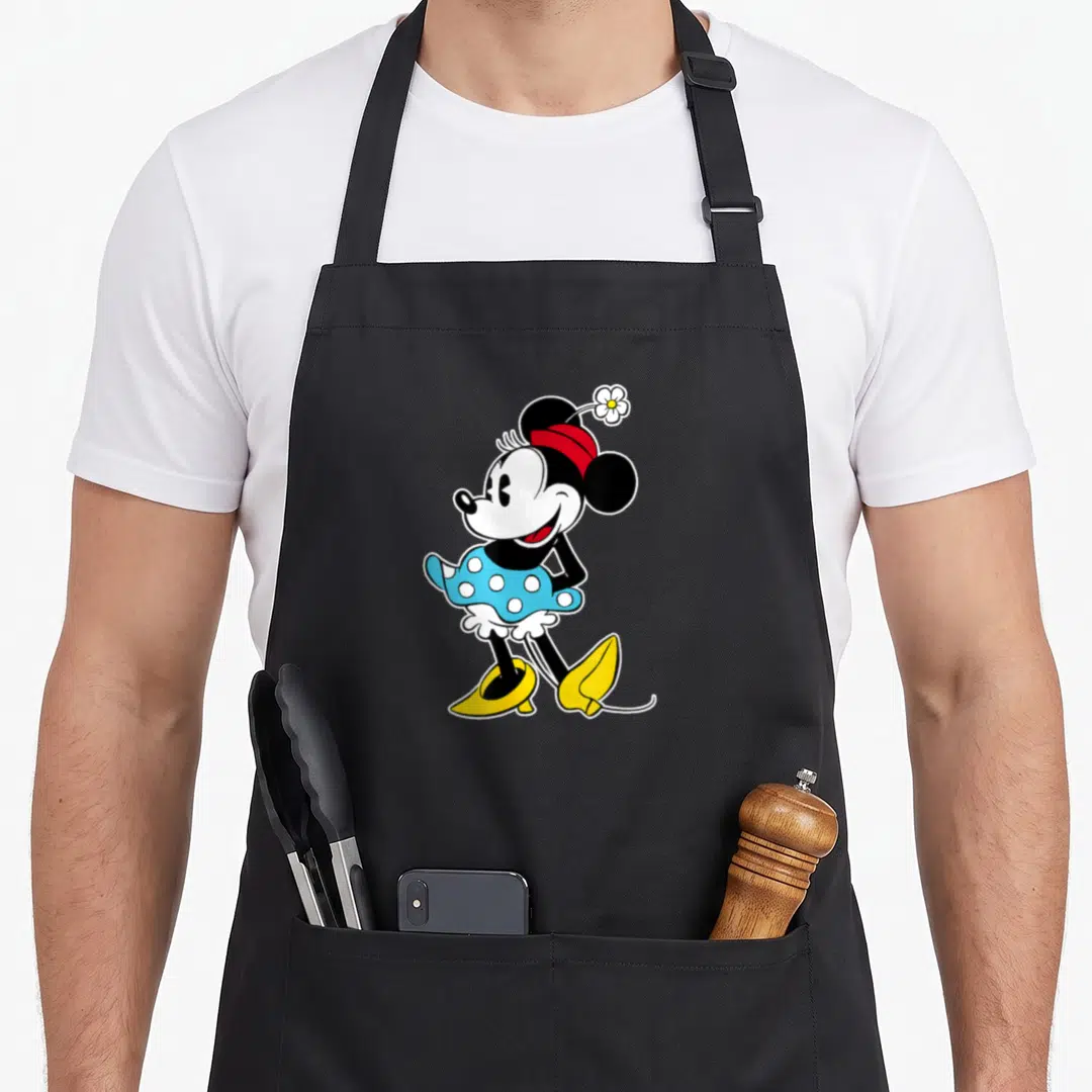 Flower mouse mom apron - Minnie Mouse standing in blue dress with white flower on red headband — shown on a black cotton canvas apron against a white background