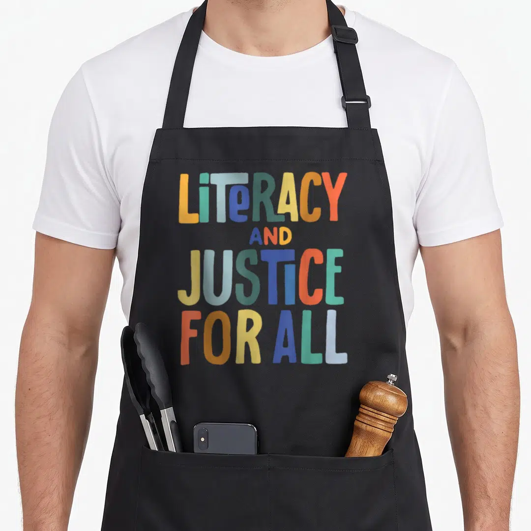 Literacy criminal justice apron - Stacked colorful text reading LITERACY AND JUSTICE FOR ALL in blocky font on grey — shown on a black cotton canvas apron against a white background