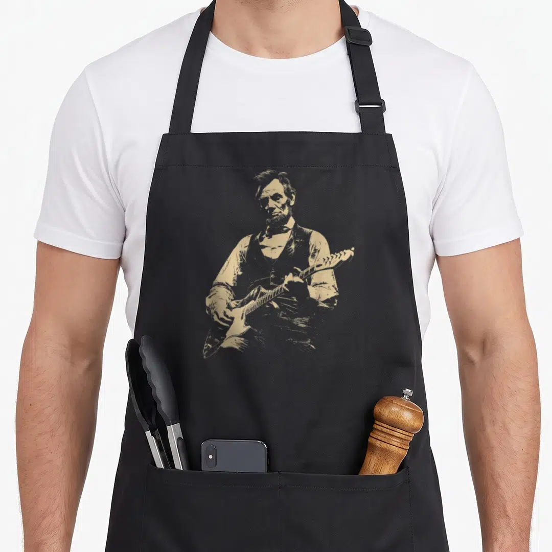 Abe playing guitar apron - Tan distressed graphic of Abraham Lincoln playing an electric guitar on canvas fabric — shown on a black cotton canvas apron against a white background