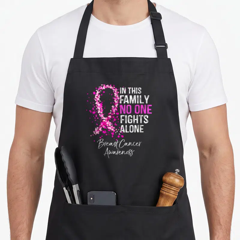 Family fight breast cancer awareness aprons - Heart-shaped pink ribbon with IN THIS FAMILY NO ONE FIGHTS ALONE text — shown on a black cotton canvas apron against a white background