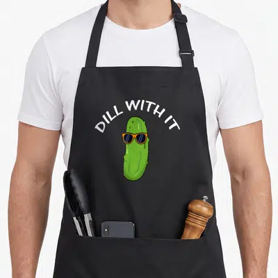 Vegetarian pickle apron - Green cartoon pickle wearing orange sunglasses under arched white text reading DILL WITH IT — shown on a black cotton canvas apron against a white background