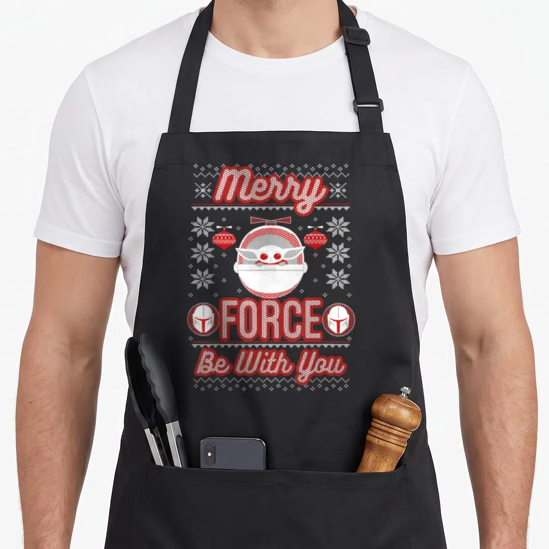 Force merry christmas apron - Grogu in pod with Merry FORCE Be With You text and Mandalorian helmets — shown on a black cotton canvas apron against a white background