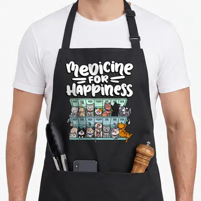 Medicine cat apron - Cartoon cats in a mint green two-tier pill organizer labeled Mon-Sun morning and evening — shown on a black cotton canvas apron against a white background