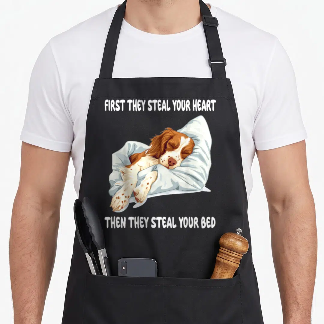 Sleeping spaniel apron - Cartoon brown and white dog sleeping on blue pillow with quote about stealing hearts and beds — shown on a black cotton canvas apron against a white background