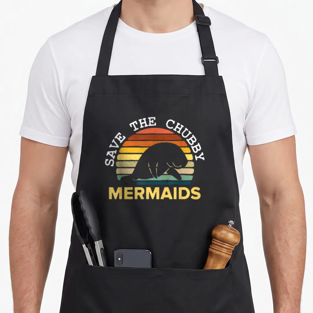 Save mermaid apron - Grey manatee silhouette against striped orange and yellow sun with text SAVE THE CHUBBY MERMAIDS — shown on a black cotton canvas apron against a white background