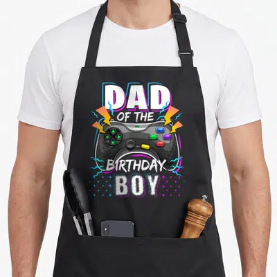 Cute video birthday apron - Black game controller flanked by yellow bolts with Dad of the Birthday Boy text — shown on a black cotton canvas apron against a white background