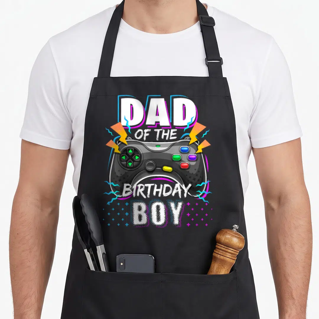 Cute video birthday apron - Black game controller flanked by yellow bolts with Dad of the Birthday Boy text — shown on a black cotton canvas apron against a white background