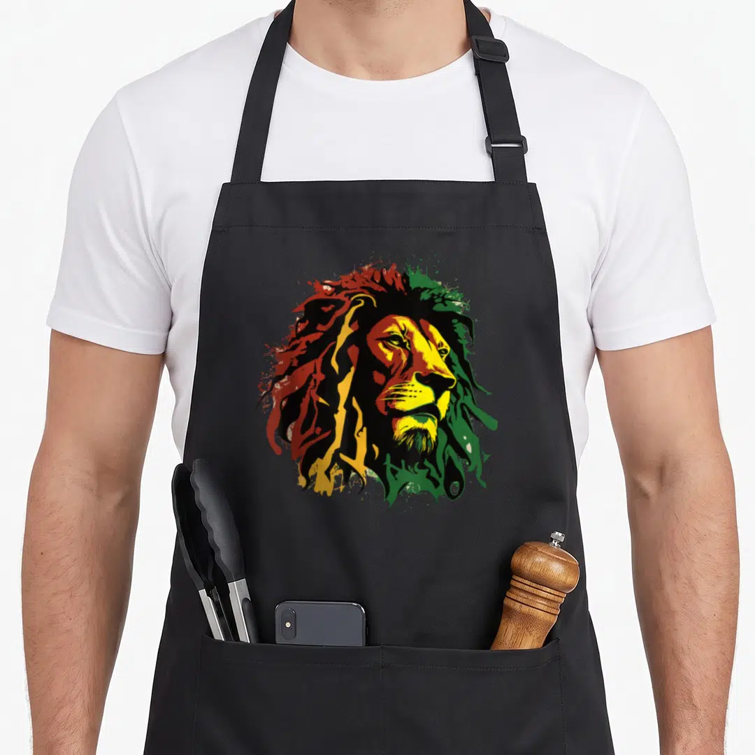 Jamaican lions apron - Stylized lion head in red yellow green black splatters cartoon art — shown on a black cotton canvas apron against a white background