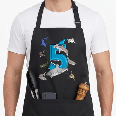 5th shark birthday apron - Blue number 5 with seven cartoon sharks swimming around it in blue, grey, and brown — shown on a black cotton canvas apron against a white background